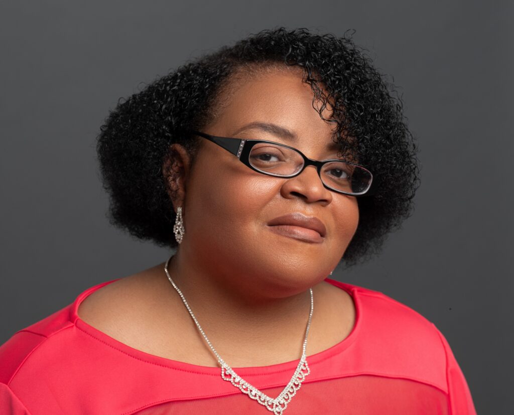 Portrait of Leisha McKinley-Beach in a coral top and glasses against a gray background.
